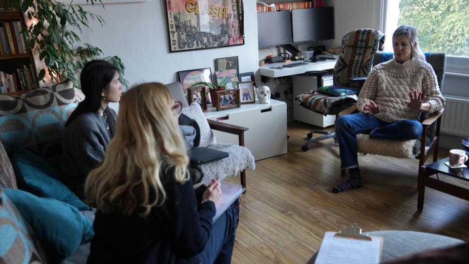 Two user researchers sit on a sofa taking notes during a conversation with a participant who is facing the camera and gesticulating as she speaks.